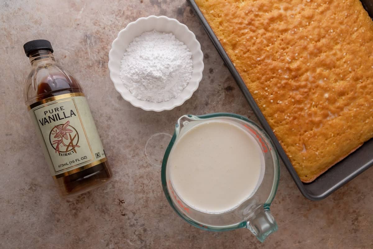 Vanilla extract, powdered sugar, and heavy cream next to a baked sponge cake for tres leches