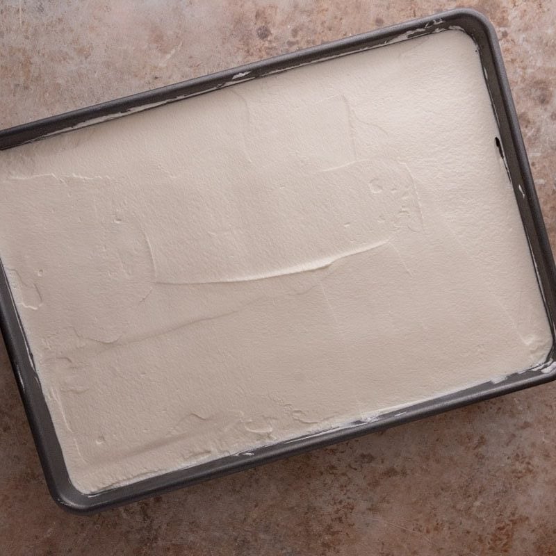 Whipped cream spread evenly over soaked tres leches cake in the pan
