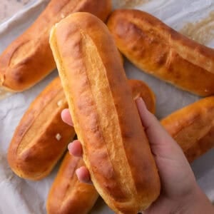 Hand holding a golden hoagie roll above a baking sheet of more homemade hoagie rolls