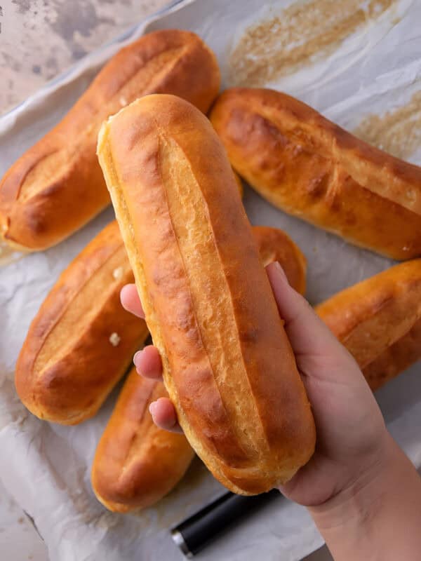 Hand holding a golden hoagie roll above a baking sheet of more homemade hoagie rolls