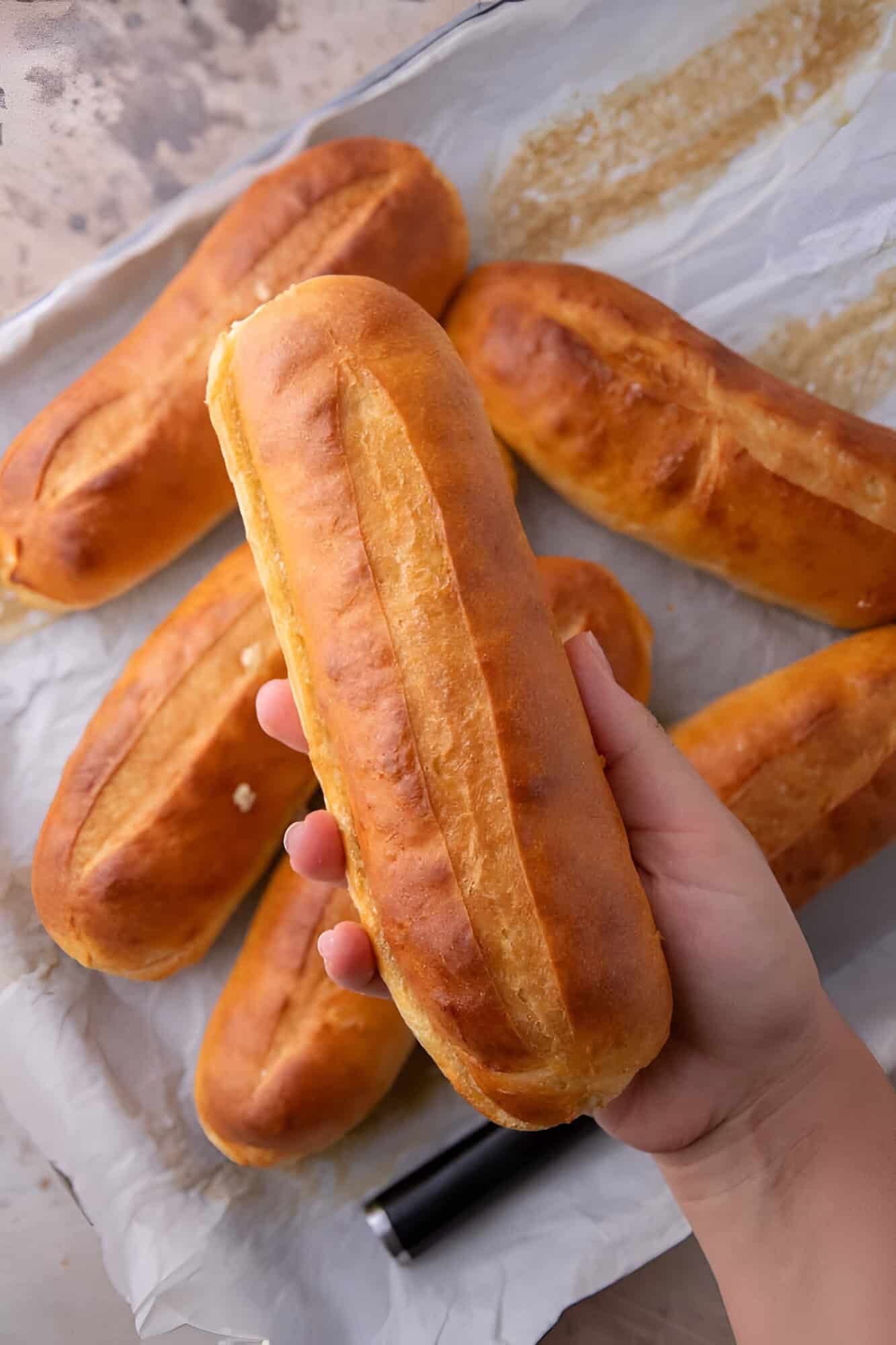 Hand holding a golden hoagie roll above a baking sheet of more homemade hoagie rolls.