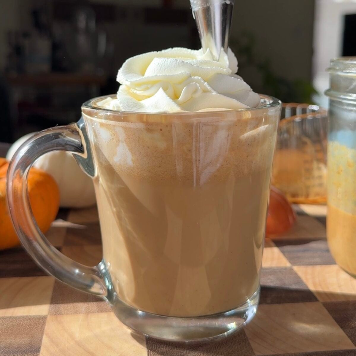 Whipped cream being piped on top of a pumpkin spice latte in a glass mug.