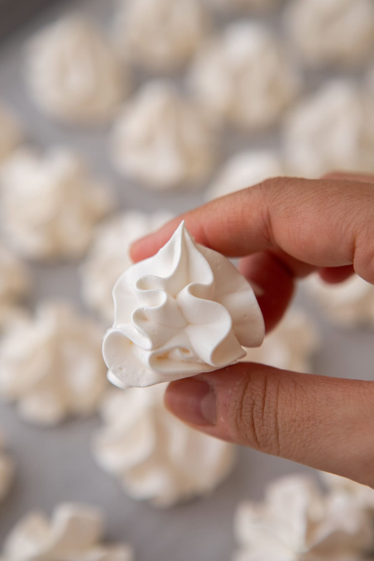 Hand holding a baked meringue cookie with a background of meringue cookies on a baking sheet.