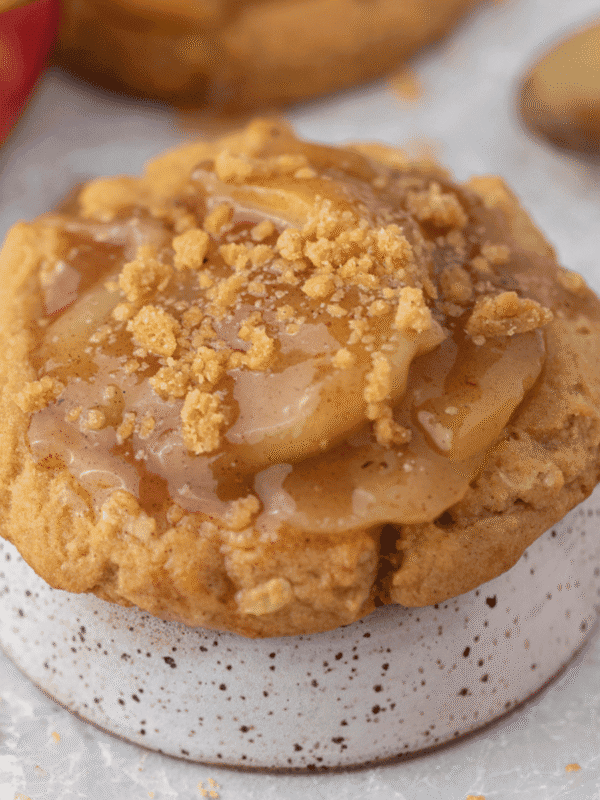 Crumbl apple pie cookie topped with glossy spiced apple pie filling and crunchy oat cookie crumbs, set on a speckled ceramic stand with a red apple behind it.