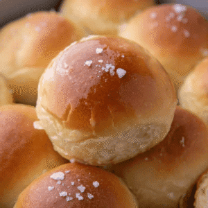 Close-up of a buttery soft dinner roll with a shiny golden top and flaky sea salt, sitting on a pan of homemade dinner rolls.