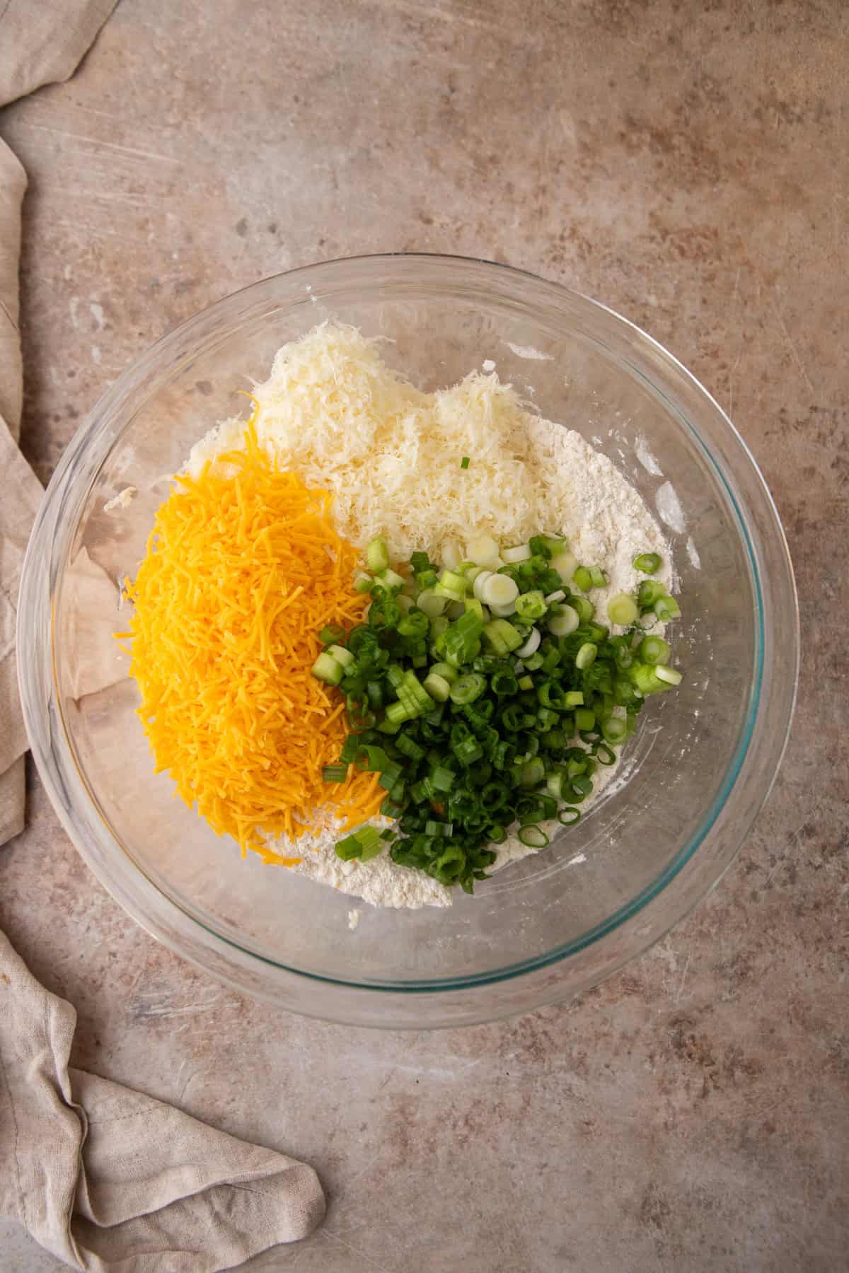 Shredded cheddar, grated parmesan, and sliced green onions added to the flour and butter mixture in a glass bowl.