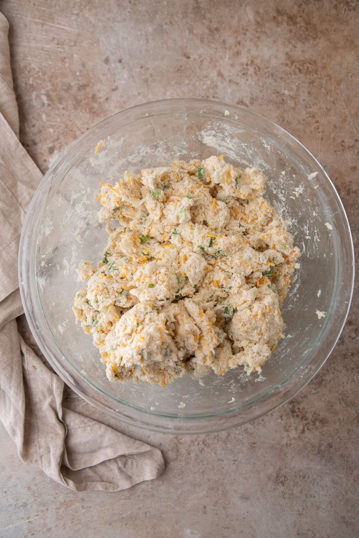 Shaggy biscuit dough with flecks of cheddar and green onion in a glass bowl.