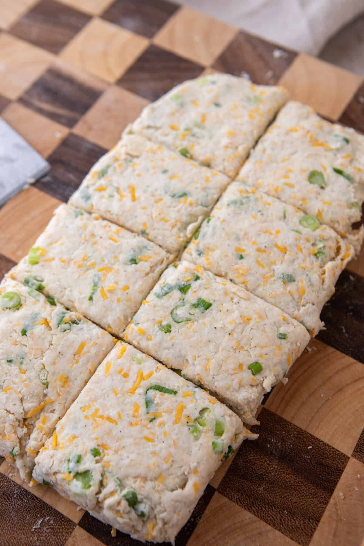 Biscuit dough cut into squares on a cutting board, with cheddar and green onion running through it.